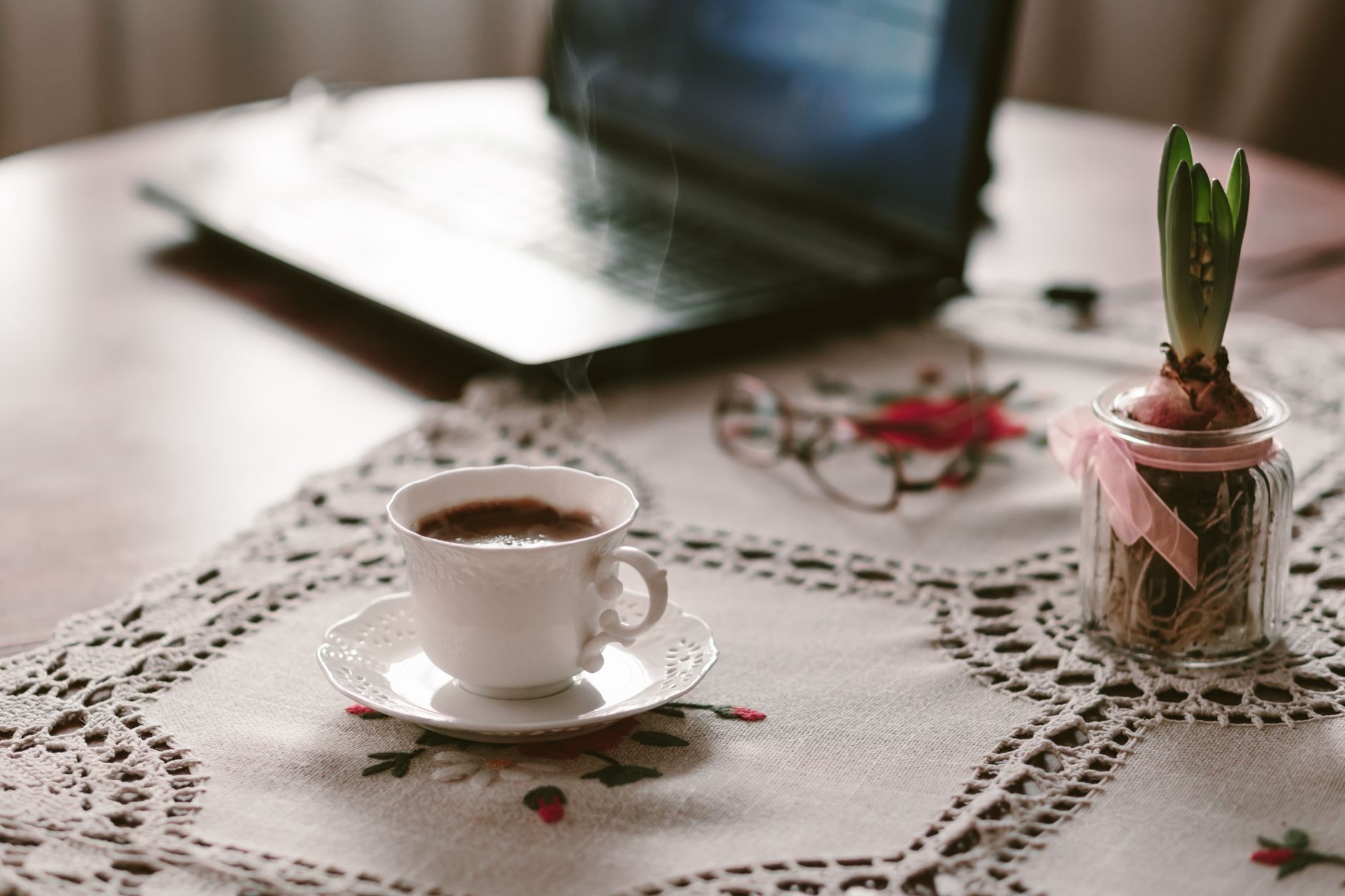 Espace de travail accueillant avec une tasse de café fumante sur une nappe en dentelle, à côté d'un ordinateur portable.